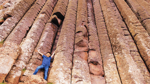 A young visitor with their hands in the air, standing on the base of a long trunk-like rock (otherwise known as the Amphitheatre) at the Giant's Causeway, Co. Antrim